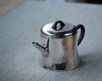 Puede incluir: Una tetera de plata con un acabado martillado y un asa negra. La tetera tiene una tapa con un pomo negro.