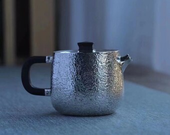 Puede incluir: Una tetera de plata con textura martillada y un asa negra. La tetera está sobre una superficie azul.