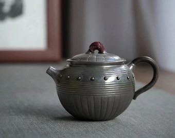 Puede incluir: Una tetera de plata con una superficie texturizada y nervada y un pomo marrón en la tapa. La tetera tiene un cuerpo redondo y un pico curvo.
