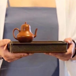 May include: A person holding a small, brown teapot on a woven tray. The teapot has a rounded body and a small spout. The tray is made of a dark brown material and has a woven pattern.