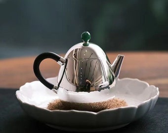 Puede incluir: Una tetera de plata con asa negra y un pomo de jade verde en la tapa. La tetera está sobre un plato de cerámica blanco con borde festoneado.