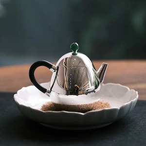 Puede incluir: Una tetera de plata con asa negra y un pomo de jade verde en la tapa. La tetera está sobre un plato de cerámica blanco con borde festoneado.