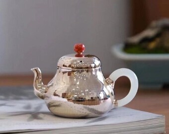 Puede incluir: Una tetera de plata con un acabado martillado y un pomo rojo en la tapa. La tetera tiene un asa de jade blanco.