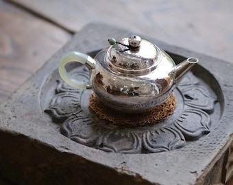 Puede incluir: Una tetera de plata con un asa de color verde jade se asienta sobre una base de piedra gris con un diseño de flor de loto tallado.