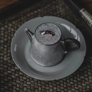 Może przedstawiać: Srebrny czajnik z fakturowaną powierzchnią i czerwonym pokrętłem na pokrywce. Czajnik stoi na białym spodeczku.