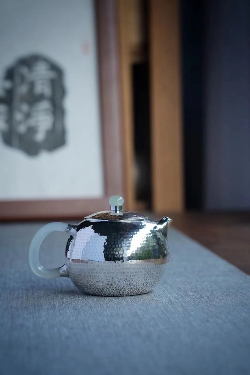 K&ouml;nnte beinhalten: Eine silberne Teekanne mit geh&auml;mmerter Oberfl&auml;che und einem jadegr&uuml;nen Knopf auf dem Deckel. Die Teekanne steht auf einer grauen Oberfl&auml;che.