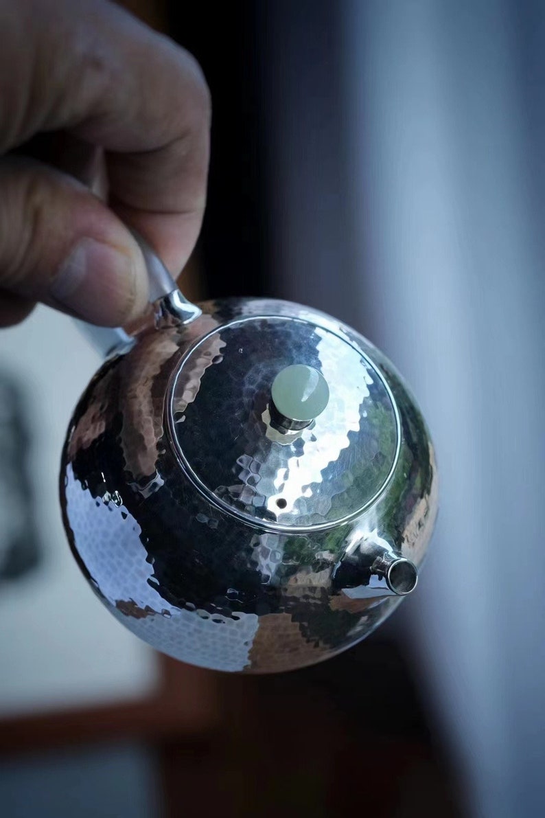 K&ouml;nnte beinhalten: Eine silberne Teekanne mit geh&auml;mmerter Oberfl&auml;che und einem jadegr&uuml;nen Knopf auf dem Deckel. Die Teekanne hat einen Ausguss und einen Griff.