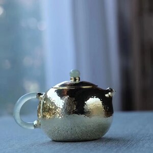 K&ouml;nnte beinhalten: Eine geh&auml;mmerte goldene Teekanne mit einem jadegr&uuml;nen Deckel und einem durchsichtigen Glasgriff. Die Teekanne steht auf einer hellgrauen Oberfl&auml;che.