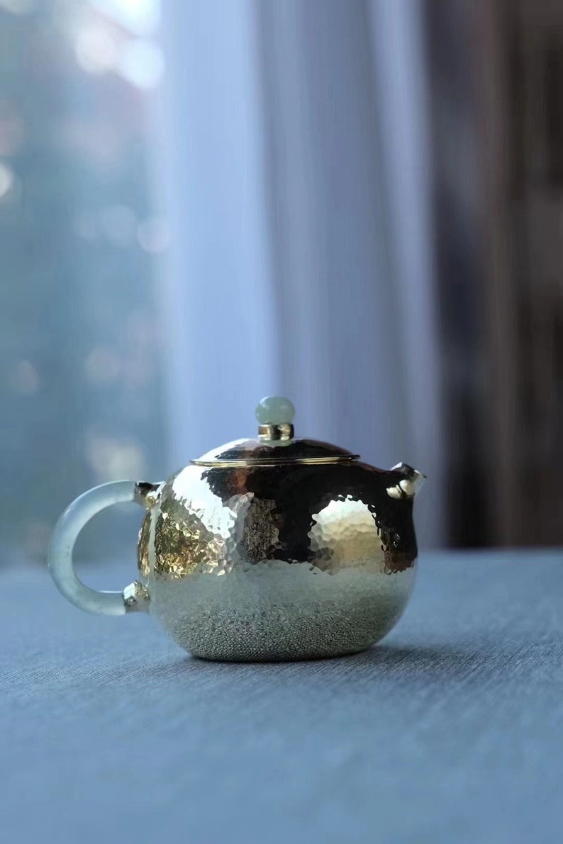 K&ouml;nnte beinhalten: Eine geh&auml;mmerte goldene Teekanne mit einem jadegr&uuml;nen Deckel und einem durchsichtigen Glasgriff. Die Teekanne steht auf einer hellgrauen Oberfl&auml;che.