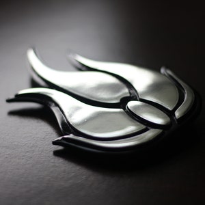 Op de afbeelding: Een zilverkleurig metalen vlamontwerp met zwarte accenten. De vlam is gestileerd en heeft een driedimensionale uitstraling.