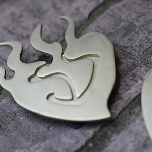 Op de afbeelding: Twee goudkleurige metalen hartvormige hangers met een gestileerd vlammenontwerp. De hangers liggen op een getextureerd grijs oppervlak.