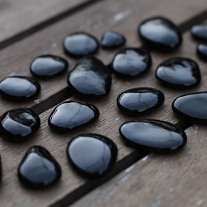 May include: A collection of smooth, black, polished stones scattered on a weathered wooden surface. The stones vary in size and shape, reflecting light and creating a glossy appearance. The arrangement suggests a natural, organic theme.