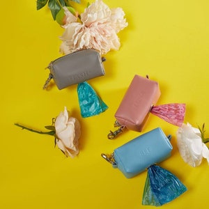 May include: A collection of dog waste bag dispensers in gray, pink, and blue, each with a matching bag. The dispensers are arranged on a bright yellow background with white and pink flowers.