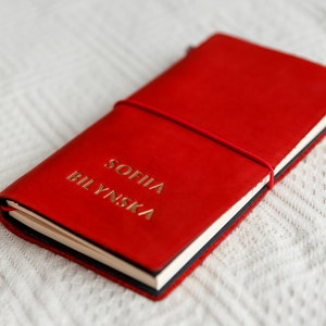May include: A red leather journal with the name "SOFIIA BILYNSKA" embossed in gold lettering. The journal is closed with a red elastic band. The notebook is lying on a white textured surface.
