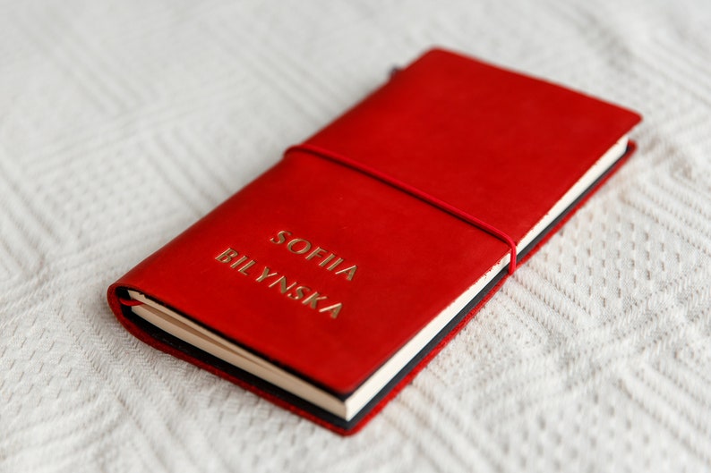 May include: A red leather journal with the name "SOFIIA BILYNSKA" embossed in gold lettering. The journal is closed with a red elastic band. The notebook is lying on a white textured surface.
