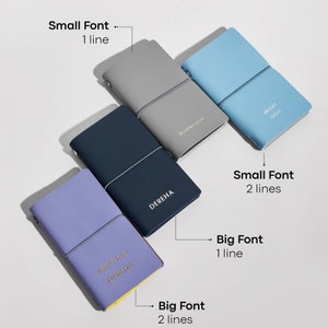 May include: Four leather-bound journals in shades of gray, blue, navy, and lavender are displayed. Each journal has a contrasting elastic band closure. The journals feature text in various fonts, including "DEREHA", "BRIGHT IDEAS", and "KRISTINA DEREHA".