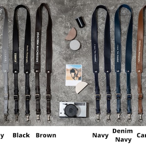 May include: Ten camera straps in different colors and materials, including grey, black, brown, navy, denim, and caramel. Each strap has a different inscription, such as "#SMILE", "Dercha Photography", "JOACHIM MAGNUSSEN", and "I Love You Grandad".