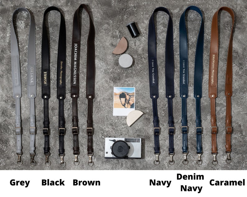 May include: Ten camera straps in different colors and materials, including grey, black, brown, navy, denim, and caramel. Each strap has a different inscription, such as "#SMILE", "Dercha Photography", "JOACHIM MAGNUSSEN", and "I Love You Grandad".