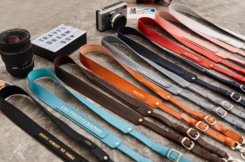 May include: A variety of leather camera straps with personalized text, including "Travelgang", "Justice", "SWJ Photography", and "Don't Forget to Smile".