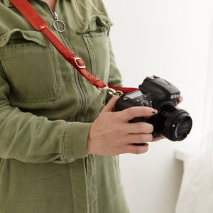May include: A person wearing a green jumpsuit holds a black Nikon camera with a red leather camera strap.