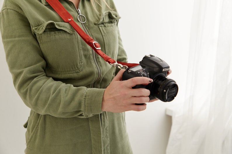 May include: A person wearing a green jumpsuit holds a black Nikon camera with a red leather camera strap.