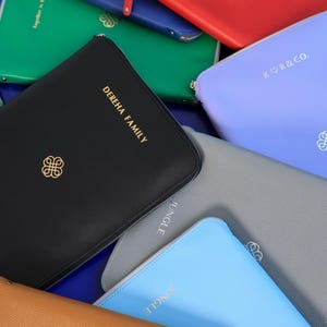 May include: A collection of colorful zippered pouches in various shades, including black, red, blue, green, and gray. Some pouches feature gold lettering with phrases like "DEREHA FAMILY" and "JUNGLE". Each pouch has a gold emblem.