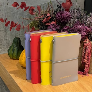 May include: A collection of colorful leather journals in purple, red, yellow, and gray, arranged on a wooden surface. The journals are secured with elastic bands and feature a minimalist design. The background includes fall foliage and gourds.