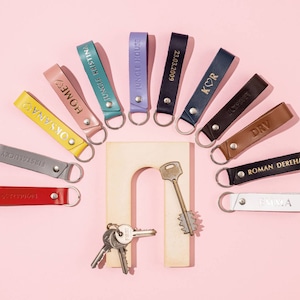 May include: A collection of colorful leather keychains with various names and phrases engraved on them. The keychains are arranged in a semi-circle on a pink background.