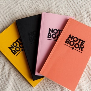 May include: Four notebooks in yellow, black, pink, and coral. Each notebook has the word "NOTE BOOK" printed in black, bold font. The notebooks are arranged in a fan shape on a white, textured surface.