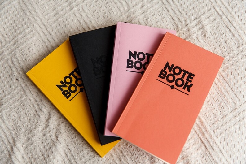 May include: Four notebooks in yellow, black, pink, and coral. Each notebook has the word "NOTE BOOK" printed in black, bold font. The notebooks are arranged in a fan shape on a white, textured surface.