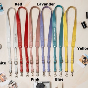 May include: Ten camera straps in different colors, including white, red, pink, lavender, blue, and yellow. Each strap has a metal buckle and a metal ring. The straps are made of leather and have different text printed on them.