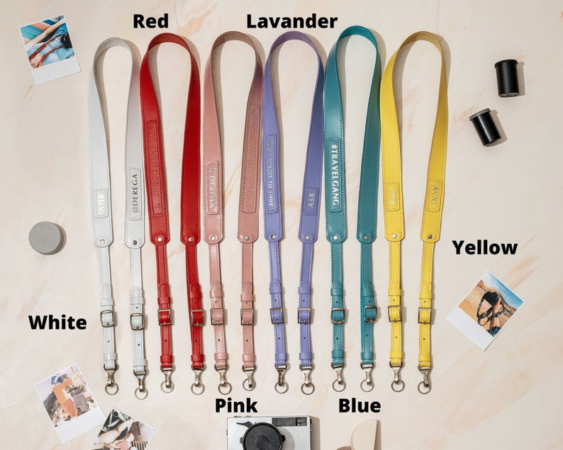 May include: Ten camera straps in different colors, including white, red, pink, lavender, blue, and yellow. Each strap has a metal buckle and a metal ring. The straps are made of leather and have different text printed on them.