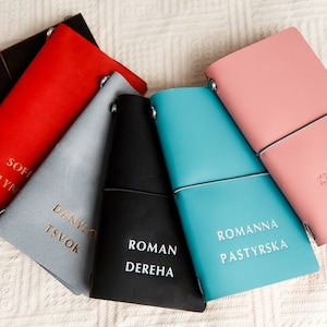 May include: A collection of leather journals in various colors, including black, red, gray, and teal, each with a name embossed in gold or white lettering. The journals are arranged in a fan shape on a white textured surface.