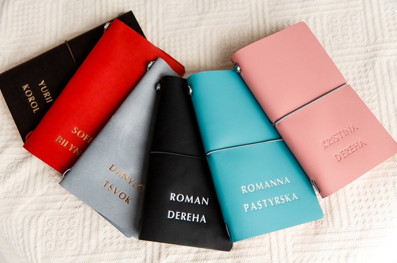 May include: A collection of leather journals in various colors, including black, red, gray, and teal, each with a name embossed in gold or white lettering. The journals are arranged in a fan shape on a white textured surface.