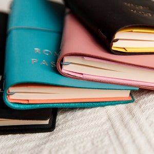 May include: A stack of leather-bound journals in various colors: black, teal, pink, and brown. The teal journal has the words "ROM PAS" embossed on the cover. The pages are visible, with different colored edges. The journals are stacked on a white, textured surface.