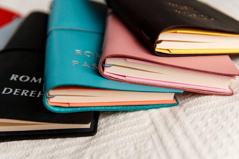 May include: A stack of leather-bound journals in various colors: black, teal, pink, and brown. The teal journal has the words "ROM PAS" embossed on the cover. The pages are visible, with different colored edges. The journals are stacked on a white, textured surface.