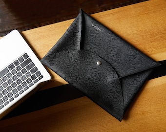May include: A black leather envelope-style clutch with a silver snap closure, resting on a wooden surface next to a laptop. The word "KRISTINA" is embossed on the clutch. The clutch has a textured surface.