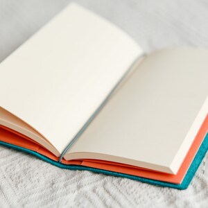 May include: An open notebook with blank, cream-colored pages. The notebook has a teal cover and orange interior pages. The notebook is lying on a white, textured surface.