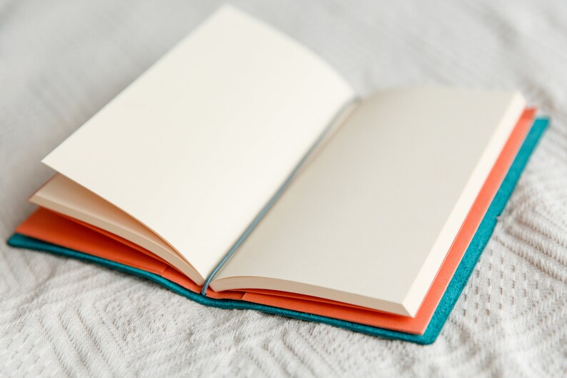 May include: An open notebook with blank, cream-colored pages. The notebook has a teal cover and orange interior pages. The notebook is lying on a white, textured surface.
