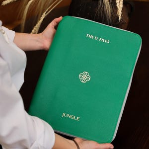 May include: A green leather-textured portfolio with the text "THE D. FILES" and "JUNGLE" in gold lettering. A gold emblem is centered on the front. The portfolio has a silver zipper and is held by a person.