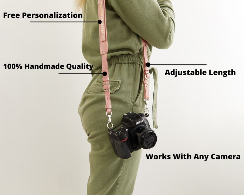 May include: A pink leather camera strap with adjustable length. The strap is worn by a person with a camera attached. The strap has the text "Free Personalization" and "100% Handmade Quality" printed on it. The text "Adjustable Length" and "Works With Any Camera" are also included in the image.