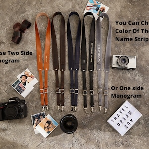 May include: Four camera straps in different colors, including brown, dark brown, navy blue, and gray. The straps are made of leather and have adjustable buckles. The straps have a gold metal name tag with the text "TRAVELGANG" and "DON'T FORGET TO SMILE". The straps are displayed on a gray textured surface with a camera, a Polaroid photo, and a sign that says "TRAVEL DREAM DO".