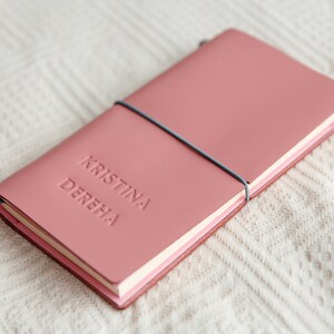May include: A dusty rose leather journal with the embossed name "KRISTINA DEREHA". The notebook is secured with a gray elastic band. The journal is lying on a white, textured surface.