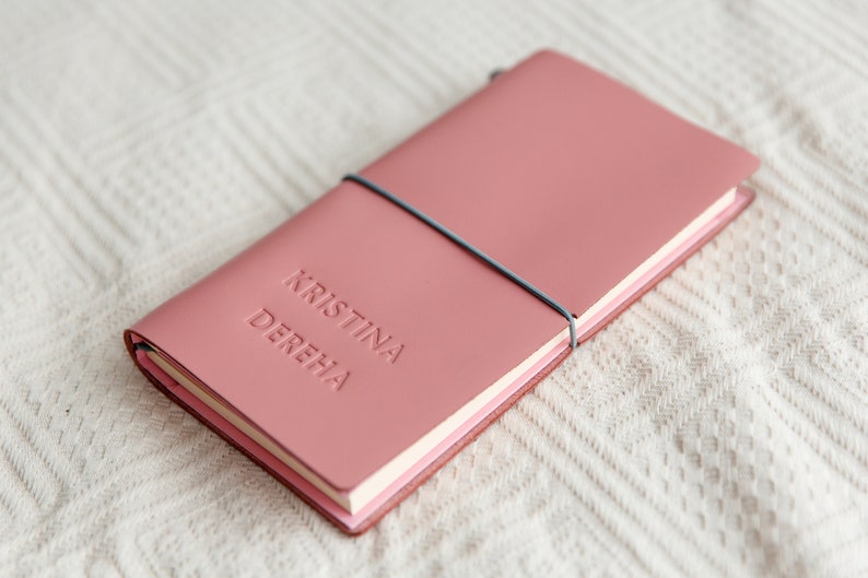 May include: A dusty rose leather journal with the embossed name "KRISTINA DEREHA". The notebook is secured with a gray elastic band. The journal is lying on a white, textured surface.