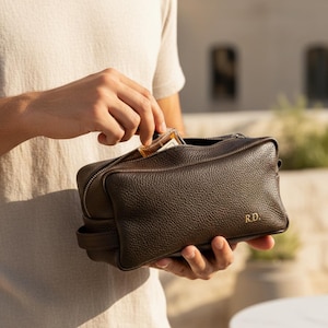 May include: A brown leather dopp kit with a zipper closure and the initials "R.D." in gold. The bag is held open, revealing a small bottle inside. The kit has a textured surface and a handle on the side.