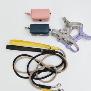 May include: A set of dog accessories including a pink and a blue leather poop bag holder, a gray and purple dog harness, and a black and beige rope leash with two clips.