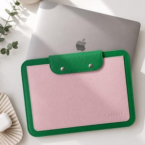 May include: A rectangular laptop sleeve featuring a vibrant green border and a soft pink center panel. The sleeve has a green flap secured by two silver metal snaps. The text JUNGLE and K.DEREHA are embossed on the pink surface of the sleeve.