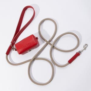 May include: A red leather dog leash with a matching red leather poop bag holder that says "SPAKKU" on it. The leash is attached to a beige rope leash with a silver clip.