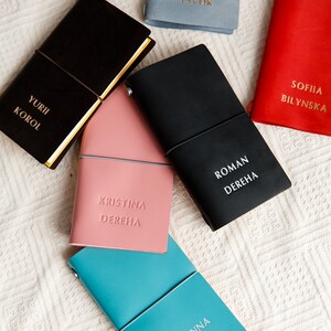 May include: A collection of personalized leather journals in various colors, including black, pink, red, and teal. Each journal features embossed names in gold or white lettering. The journals are closed with an elastic band.