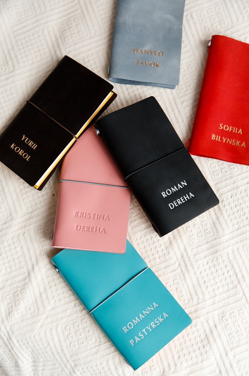 May include: A collection of personalized leather journals in various colors, including black, pink, red, and teal. Each journal features embossed names in gold or white lettering. The journals are closed with an elastic band.